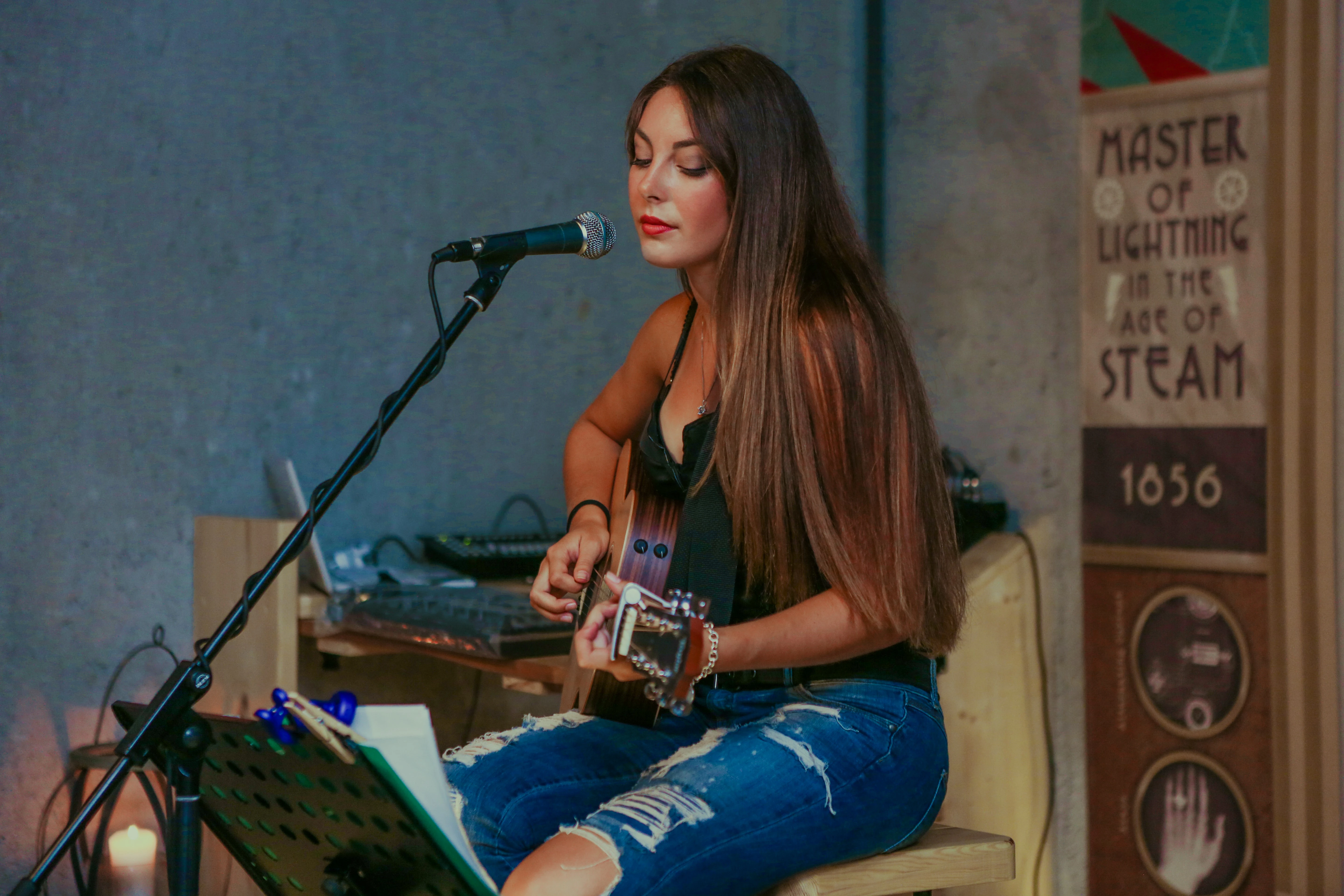 Chica joven con pelo largo tocando la guitarra delante de un micro mirando a un partitura dentro de una sala de grabación